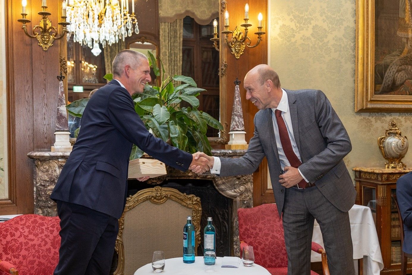 Dr. Dahmen begrüßt Henning Dehler, Geschäftsführer von Meggle bei den Kronberger Kamingesprächen 2024. Man steht vor einem Kamin. Zwischen den beiden befindet sich ein niedriger Tisch mit Wassergläsern und Wasserflaschen. Der Raum, in denen sich die Personen befinden hat einen leicht barocken Stil. Kronberger Kamingespräche 2024. Podiumsdiskussion.