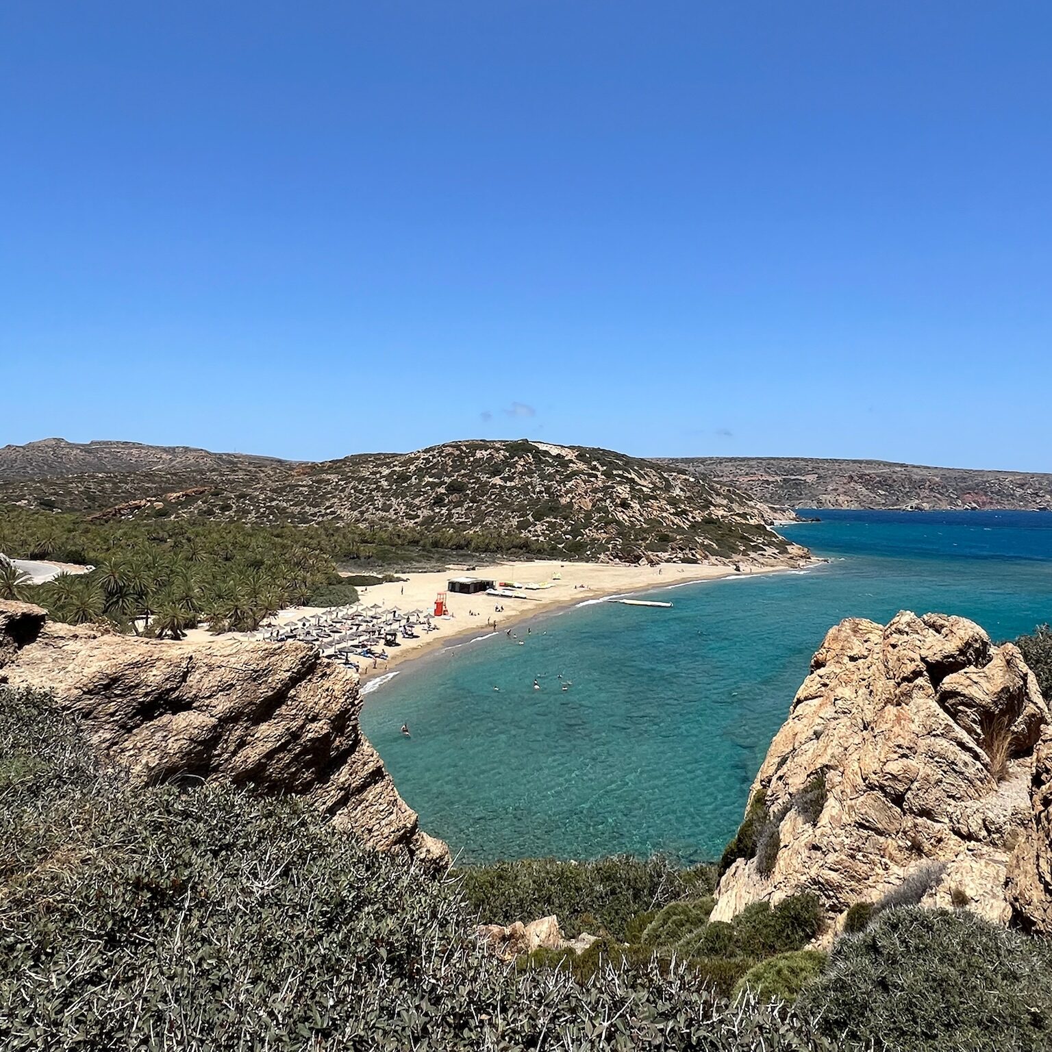 Eine Bucht in Kreta. Sandstrand mit aufgestellten Sonnenliegen. Perspektivisch schaut man von Felgen hervor.